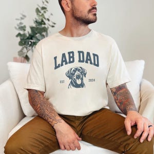 May include: A person wearing a cream colored t-shirt with the text "Lab Dad Est 2024" and a black Labrador Retriever silhouette. The shirt has a vintage collegiate style font.