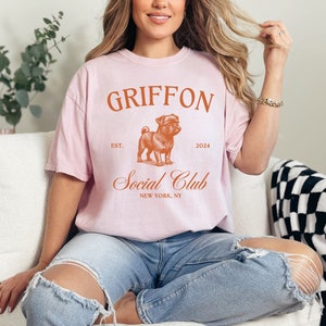 May include: A pink t-shirt with the text "GRIFFON Social Club" in orange. The text "EST. 2024" is also in orange. A cartoon image of a pug is in the center of the shirt.