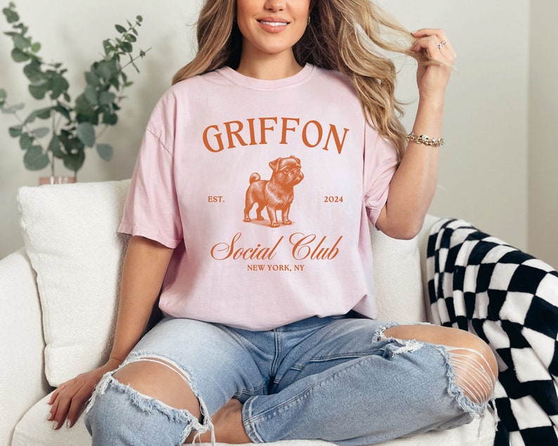 May include: A pink t-shirt with the text "GRIFFON Social Club" in orange. The text "EST. 2024" is also in orange. A cartoon image of a pug is in the center of the shirt.