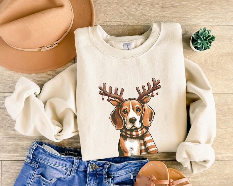May include: A beige sweatshirt with a cartoon image of a beagle dog wearing a red and brown reindeer antler headband with red berries. The dog is wearing a striped scarf and has a brown nose.