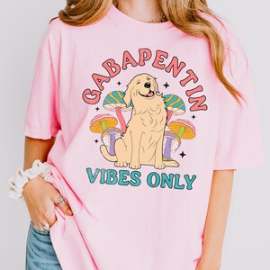 K&ouml;nnte beinhalten: Pinkes T-Shirt mit einem Grafikdruck eines Golden Retriever Hundes, der vor bunten Pilzen sitzt. Der Text "Gabapentin Vibes Only" ist auf dem Shirt aufgedruckt.