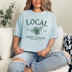 May include: A light blue t-shirt with a green graphic of a bee and the text "LOCAL EST. 2024 HONEY DEALER SAN FRANCISCO, CA"