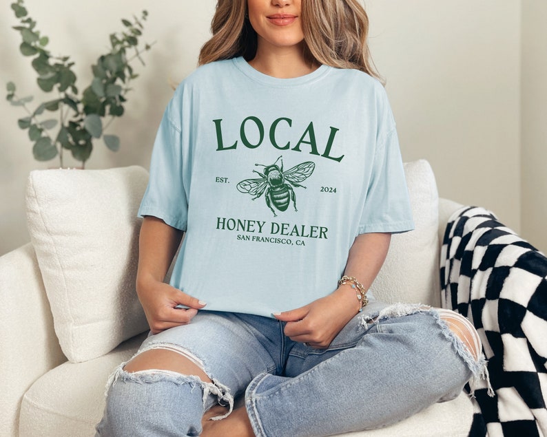 May include: A light blue t-shirt with a green graphic of a bee and the text "LOCAL EST. 2024 HONEY DEALER SAN FRANCISCO, CA"