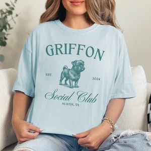 May include: Light blue t-shirt with a green graphic of a pug wearing glasses. The graphic says "Griffon Social Club Est. 2024 Austin, TX"