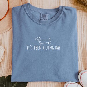May include: A blue t-shirt featuring the text "It's been a long day" and a white dachshund dog illustration. The shirt is made of a soft material and is a comfortable fit.