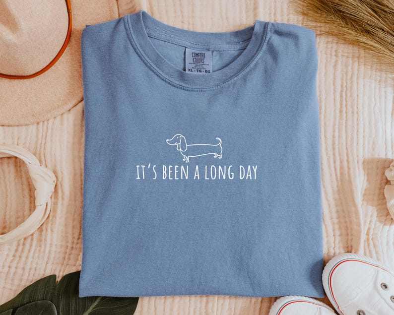 May include: A blue t-shirt featuring the text "It's been a long day" and a white dachshund dog illustration. The shirt is made of a soft material and is a comfortable fit.