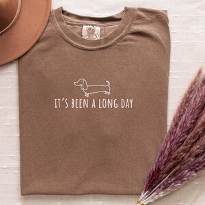 May include: A brown t-shirt with the phrase "IT'S BEEN A LONG DAY" and a white dachshund illustration. A brown hat and a bundle of dried purple flowers are also in the image.