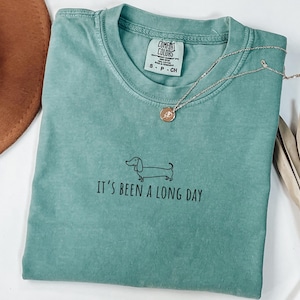 May include: A teal Comfort Colors t-shirt with the text "It's been a long day" and a dachshund illustration. A gold necklace with a pendant is draped on the shirt. A brown hat is in the upper left corner.
