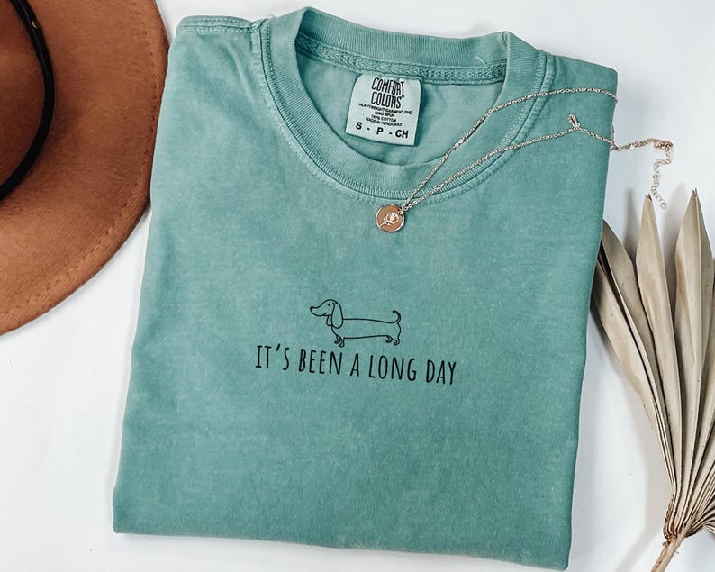 May include: A teal Comfort Colors t-shirt with the text "It's been a long day" and a dachshund illustration. A gold necklace with a pendant is draped on the shirt. A brown hat is in the upper left corner.