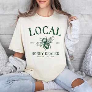 May include: A cream coloured t-shirt with a green graphic of a bee and the text "LOCAL EST. 2024 HONEY DEALER CUSTOM LOCATION"