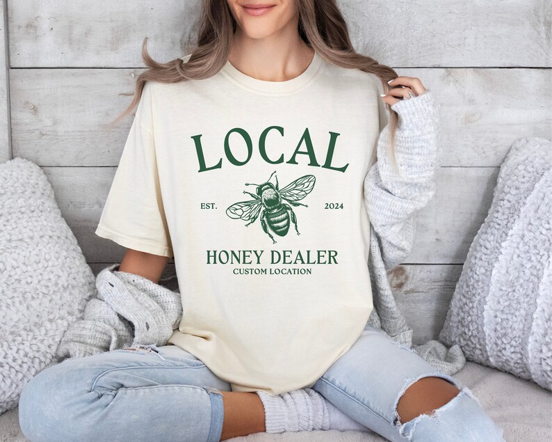 May include: A cream coloured t-shirt with a green graphic of a bee and the text "LOCAL EST. 2024 HONEY DEALER CUSTOM LOCATION"