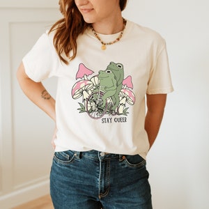 May include: A white t-shirt with a graphic of two frogs riding a bicycle through pink mushrooms. The text "STAY QUEER" is printed below the image.