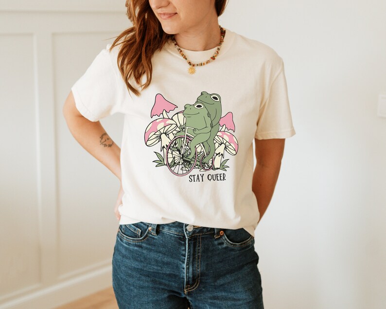 May include: A white t-shirt with a graphic of two frogs riding a bicycle through pink mushrooms. The text "STAY QUEER" is printed below the image.