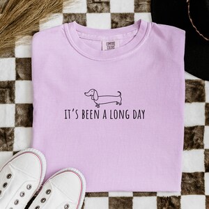 May include: A lilac sweatshirt featuring a dachshund illustration and the phrase "IT'S BEEN A LONG DAY". The image includes white trainers and a checkered pattern background.