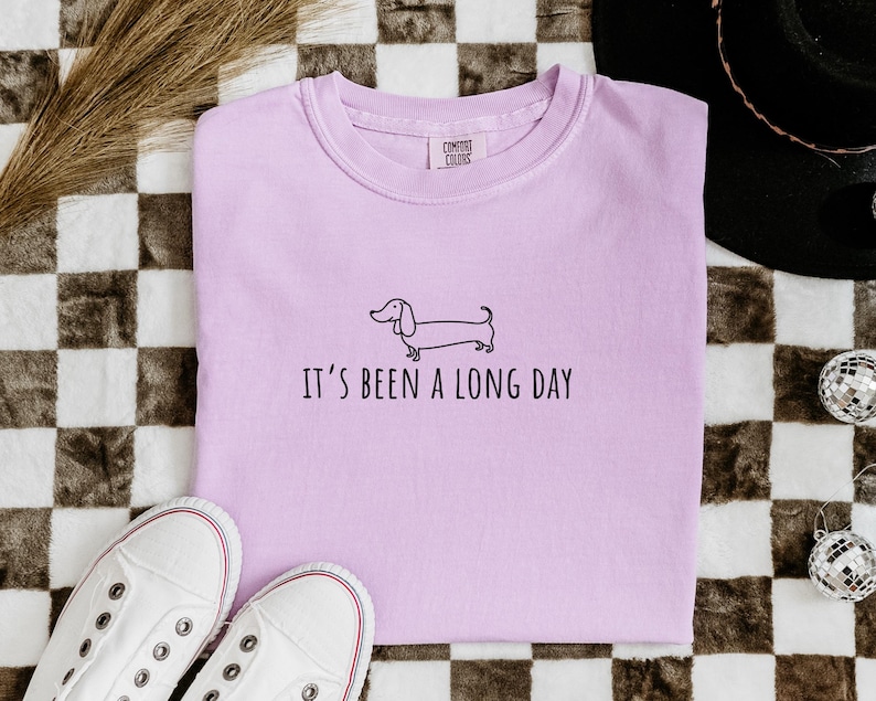 May include: A lilac sweatshirt featuring a dachshund illustration and the phrase "IT'S BEEN A LONG DAY". The image includes white trainers and a checkered pattern background.