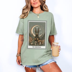 May include: A green t-shirt with a graphic of a Doberman Pinscher sitting on a crescent moon with mushrooms and stars. The text "The Doberman" is printed below the graphic.