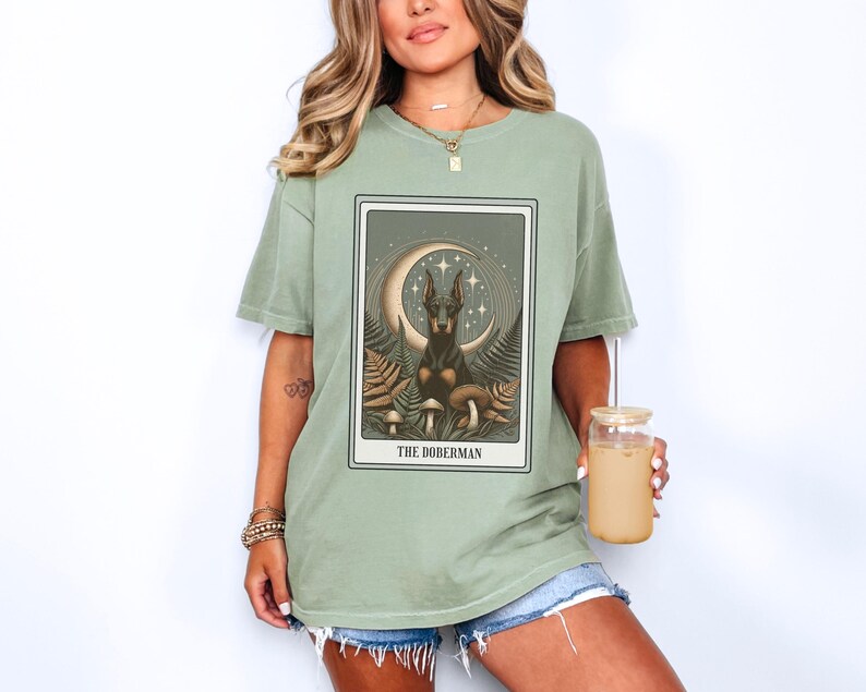 May include: A green t-shirt with a graphic of a Doberman Pinscher sitting on a crescent moon with mushrooms and stars. The text "The Doberman" is printed below the graphic.