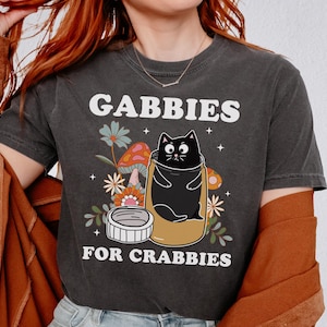 May include: A black t-shirt with a black cat in a jar with flowers and mushrooms. The text on the shirt reads "Gabbie's for Crabbies".