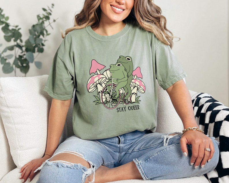 May include: A green t-shirt with a graphic of two frogs riding a bicycle with mushrooms in the background. The text "STAY QUEER" is printed below the frogs.