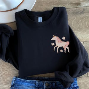 May include: Black sweatshirt with a pink and gold unicorn graphic on the left chest. The unicorn is surrounded by small white flowers.