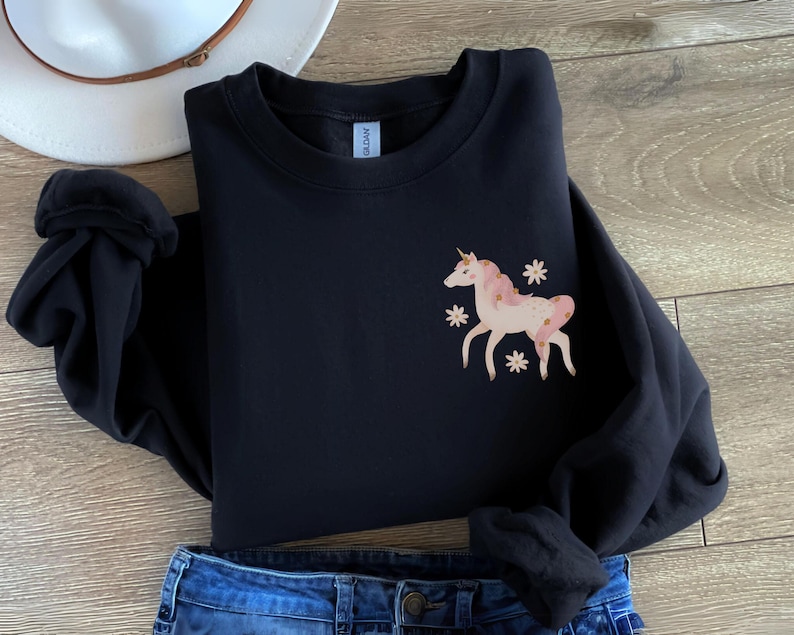 May include: Black sweatshirt with a pink and gold unicorn graphic on the left chest. The unicorn is surrounded by small white flowers.