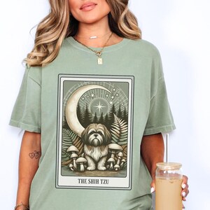 May include: Sage green t-shirt with a graphic print of a dog in a forest setting with a crescent moon and stars. The text "THE SHIH TZU" is printed below the image.
