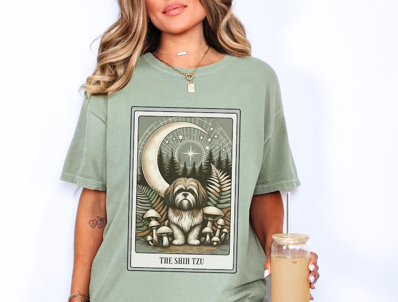 May include: Sage green t-shirt with a graphic print of a dog in a forest setting with a crescent moon and stars. The text "THE SHIH TZU" is printed below the image.