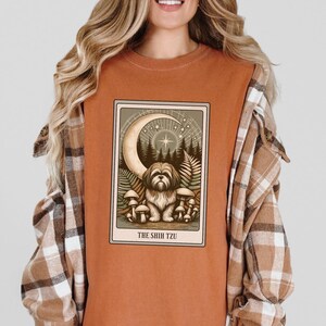 May include: A rust-colored t-shirt with a graphic of a tarot card featuring a Shih Tzu dog sitting in a forest with a crescent moon and stars above. The card reads "The Shih Tzu".