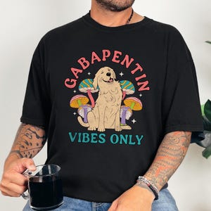 K&ouml;nnte beinhalten: Schwarzes T-Shirt mit einem Grafikdruck eines Hundes, der vor Pilzen sitzt. Der Text "Gabapentin Vibes Only" ist auf dem Shirt aufgedruckt.