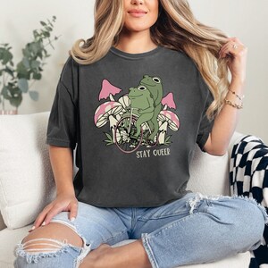 May include: A dark gray t-shirt with a graphic of two frogs riding a bicycle through a field of mushrooms. The text "STAY QUEER" is printed below the image.