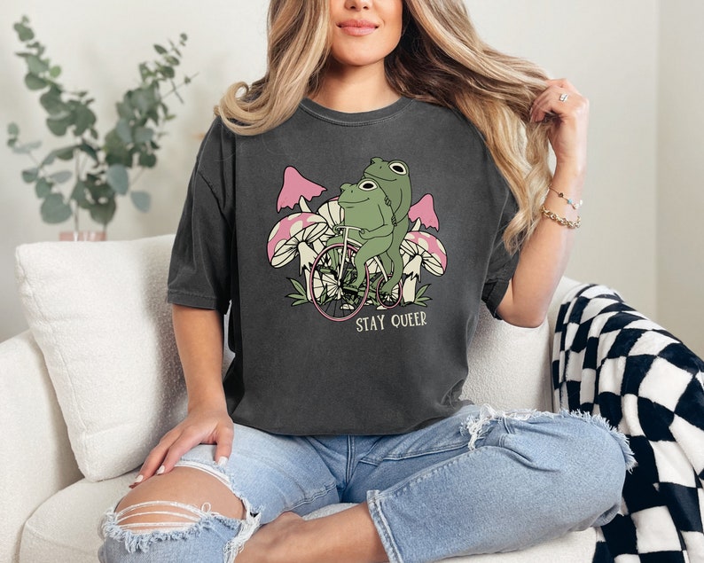 May include: A dark gray t-shirt with a graphic of two frogs riding a bicycle through a field of mushrooms. The text "STAY QUEER" is printed below the image.