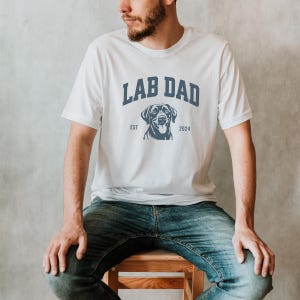 May include: White t-shirt with a blue graphic that says "Lab Dad" with a black Labrador Retriever silhouette and the text "EST 2024".