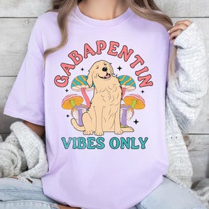 K&ouml;nnte beinhalten: Lilafarbenes T-Shirt mit einem Aufdruck eines Golden Retrievers, der vor Pilzen sitzt. Der Text "Gabapentin Vibes Only" ist unter dem Hund gedruckt.
