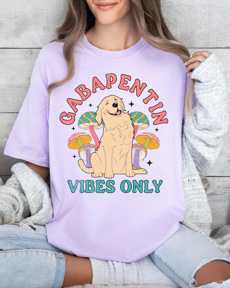 K&ouml;nnte beinhalten: Lilafarbenes T-Shirt mit einem Aufdruck eines Golden Retrievers, der vor Pilzen sitzt. Der Text "Gabapentin Vibes Only" ist unter dem Hund gedruckt.