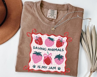 May include: A brown Comfort Colors t-shirt featuring a strawberry graphic and the text "SAVING ANIMALS IS MY JAM." The design includes pink and red strawberries on a checkered pattern, framed with red ribbons.