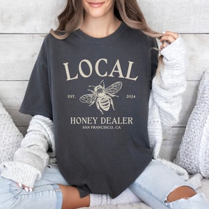 May include: A dark grey t-shirt with the text "LOCAL" in white, a bee illustration, and the text "HONEY DEALER SAN FRANCISCO, CA" in white. The text "EST. 2024" is also in white.