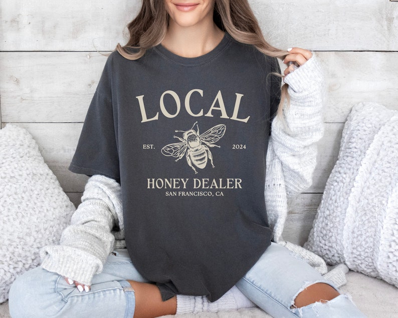 May include: A dark grey t-shirt with the text "LOCAL" in white, a bee illustration, and the text "HONEY DEALER SAN FRANCISCO, CA" in white. The text "EST. 2024" is also in white.