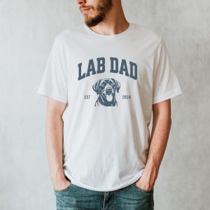 May include: White t-shirt with the text "Lab Dad" in blue, featuring a black Labrador Retriever silhouette and the text "Est 2024".
