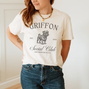 May include: A white t-shirt with a black and white illustration of a pug. The shirt has the text "GRIFFON EST. 2024 Social Club SAN FRANCISCO, CA" printed on it.