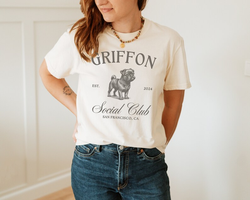 May include: A white t-shirt with a black and white illustration of a pug. The shirt has the text "GRIFFON EST. 2024 Social Club SAN FRANCISCO, CA" printed on it.