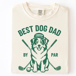 May include: Cream-colored t-shirt with a green graphic. The graphic features the text "BEST DOG DAD" above a dog wearing a hat that says "COOPER". Below the dog are the words "BY PAR" and crossed golf clubs and a golf ball.