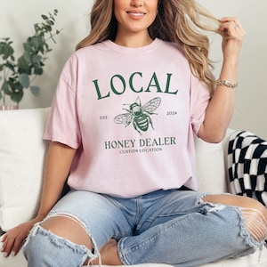 May include: A pink t-shirt with a green bee graphic and the text "LOCAL EST. 2024 HONEY DEALER CUSTOM LOCATION".