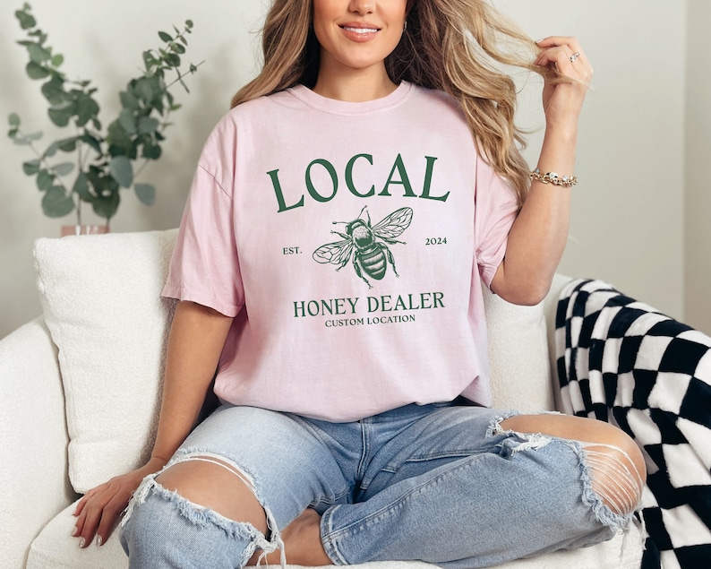 May include: A pink t-shirt with a green bee graphic and the text "LOCAL EST. 2024 HONEY DEALER CUSTOM LOCATION".