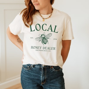 May include: A white t-shirt with a green graphic of a bee and the text "LOCAL EST. 2024 HONEY DEALER SAN FRANCISCO, CA"