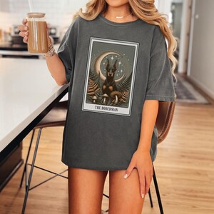 May include: A dark gray oversized t-shirt with a graphic print of a Doberman Pinscher dog in a mystical setting with a crescent moon, stars, and mushrooms. The text "THE DOBERMAN" is printed below the image.