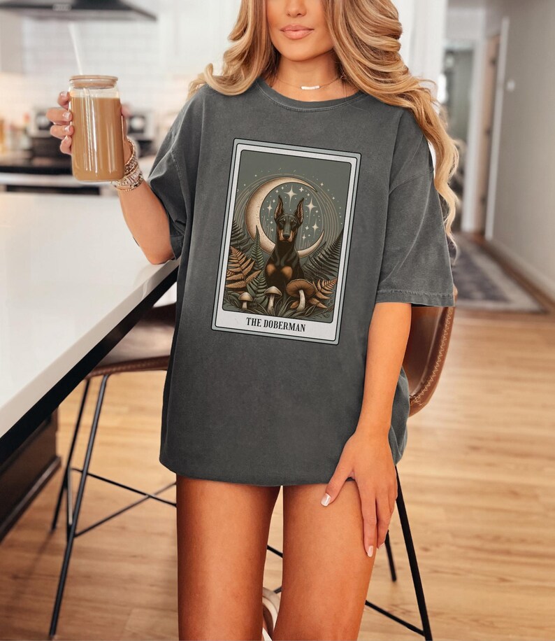 May include: A dark gray oversized t-shirt with a graphic print of a Doberman Pinscher dog in a mystical setting with a crescent moon, stars, and mushrooms. The text "THE DOBERMAN" is printed below the image.