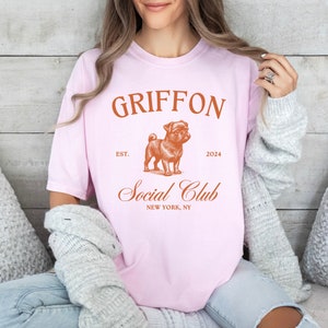 May include: Pink t-shirt with a brown graphic of a Griffon dog and the text "Griffon Social Club Est. 2024 New York, NY"