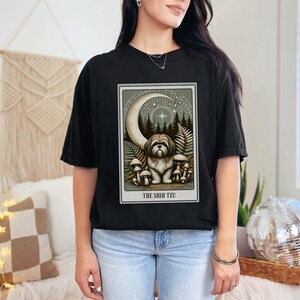 May include: Black t-shirt with a graphic of a Shih Tzu dog sitting in a forest with a crescent moon and stars. The text "THE SHIH TZU" is below the image.