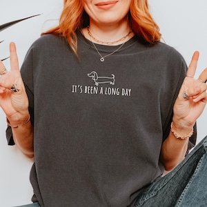 May include: Charcoal grey t-shirt featuring the phrase "IT'S BEEN A LONG DAY" and a white dachshund outline. The top has a casual style, suitable for everyday wear.