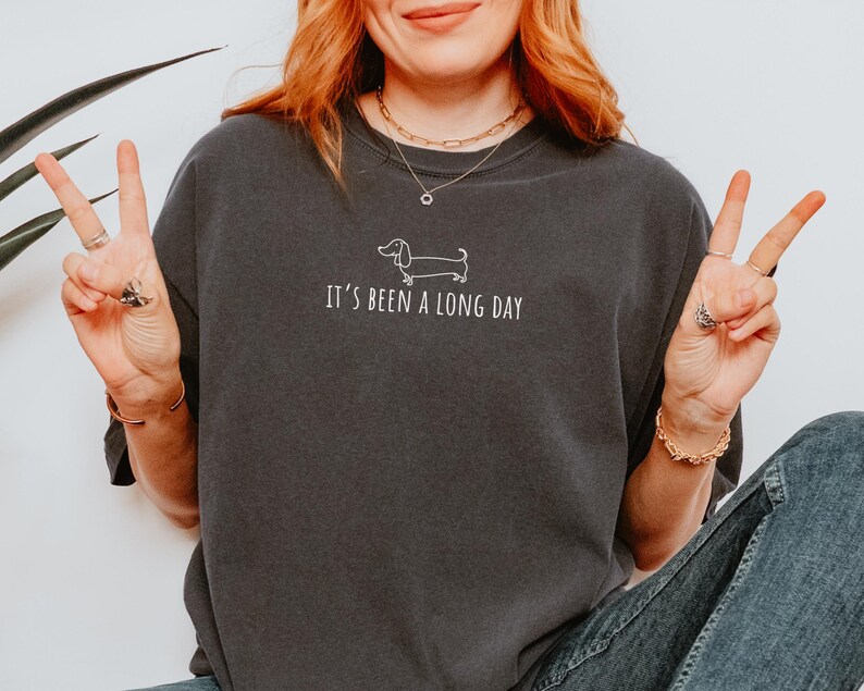 May include: Charcoal grey t-shirt featuring the phrase "IT'S BEEN A LONG DAY" and a white dachshund outline. The top has a casual style, suitable for everyday wear.